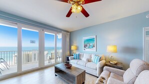 Ein geräumiges Wohnzimmer mit großem Fenster zum Meer, einem Deckenventilator, einem weißen Sofa, einem beigefarbenen Sofa und einem Couchtisch.