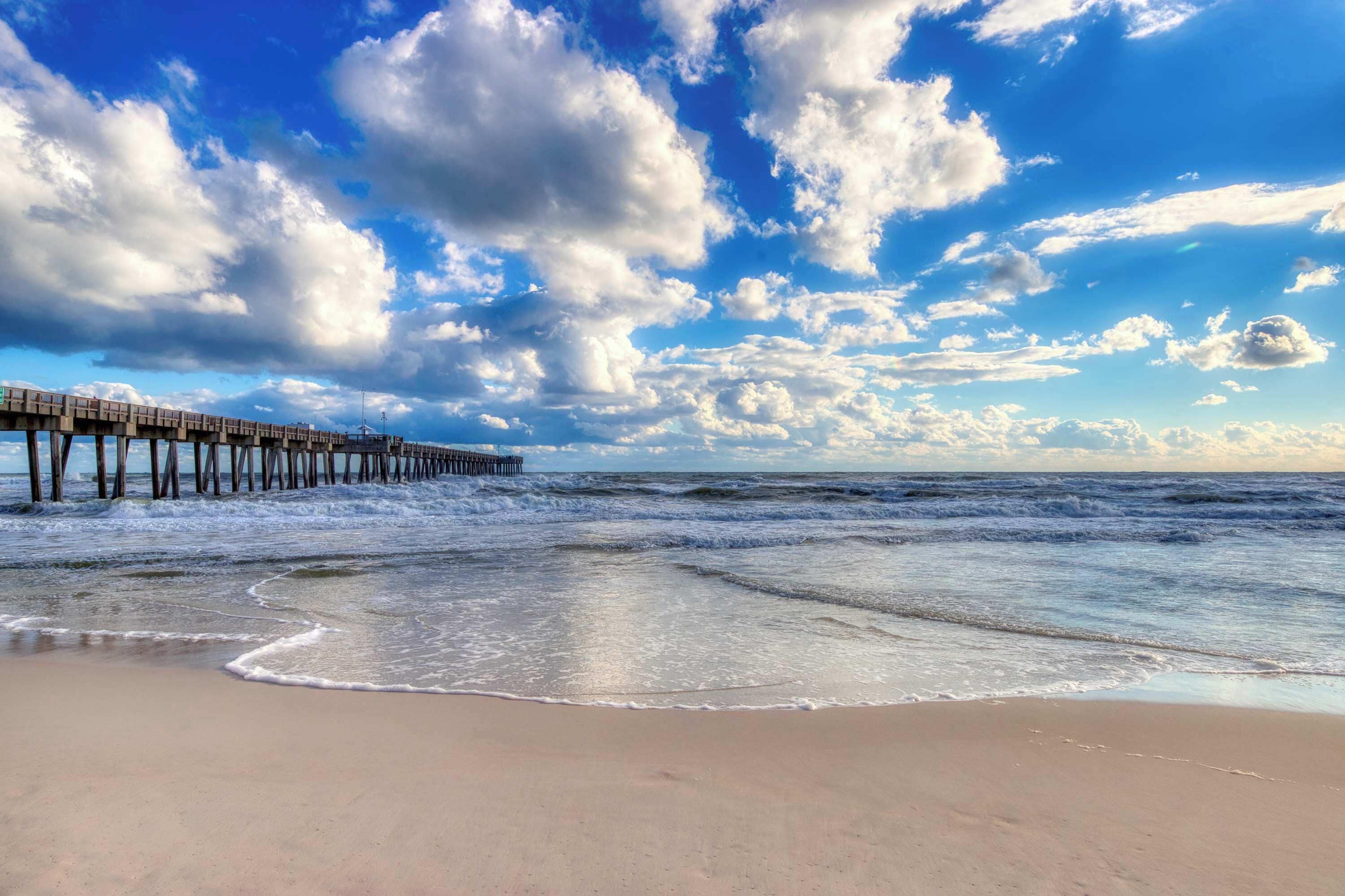 A beach with a wooden pier extending into the ocean, waves gently lapping at the shore, and a partly cloudy sky.