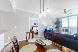 Uma sala de jantar com mesa de madeira, cadeiras e uma tigela de frutas. Há um ventilador de teto e luminárias pendentes acima da mesa.