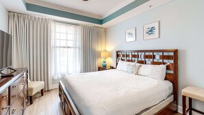 Une chambre à coucher bien rangée, avec un grand lit, une tête de lit en bois, une table de chevet avec une lampe, un téléviseur et un petit tabouret.