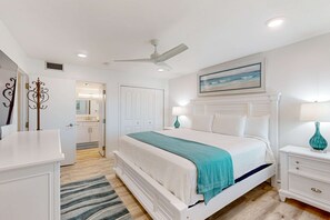 Un dormitorio con una cama, mesitas de noche, un ventilador de techo, un cuadro y una puerta que da a un baño.