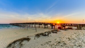 Une plage avec une jetée en bois qui s’avance dans l’océan, un ciel dégagé et le soleil se couchant à l’horizon.
