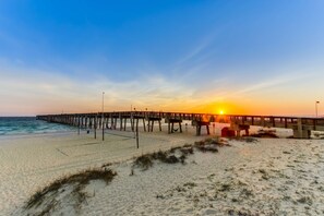 Ein Strand mit einem hölzernen Steg, der sich ins Meer erstreckt, bewölkter Himmel und ein Sonnenuntergang am Horizont.