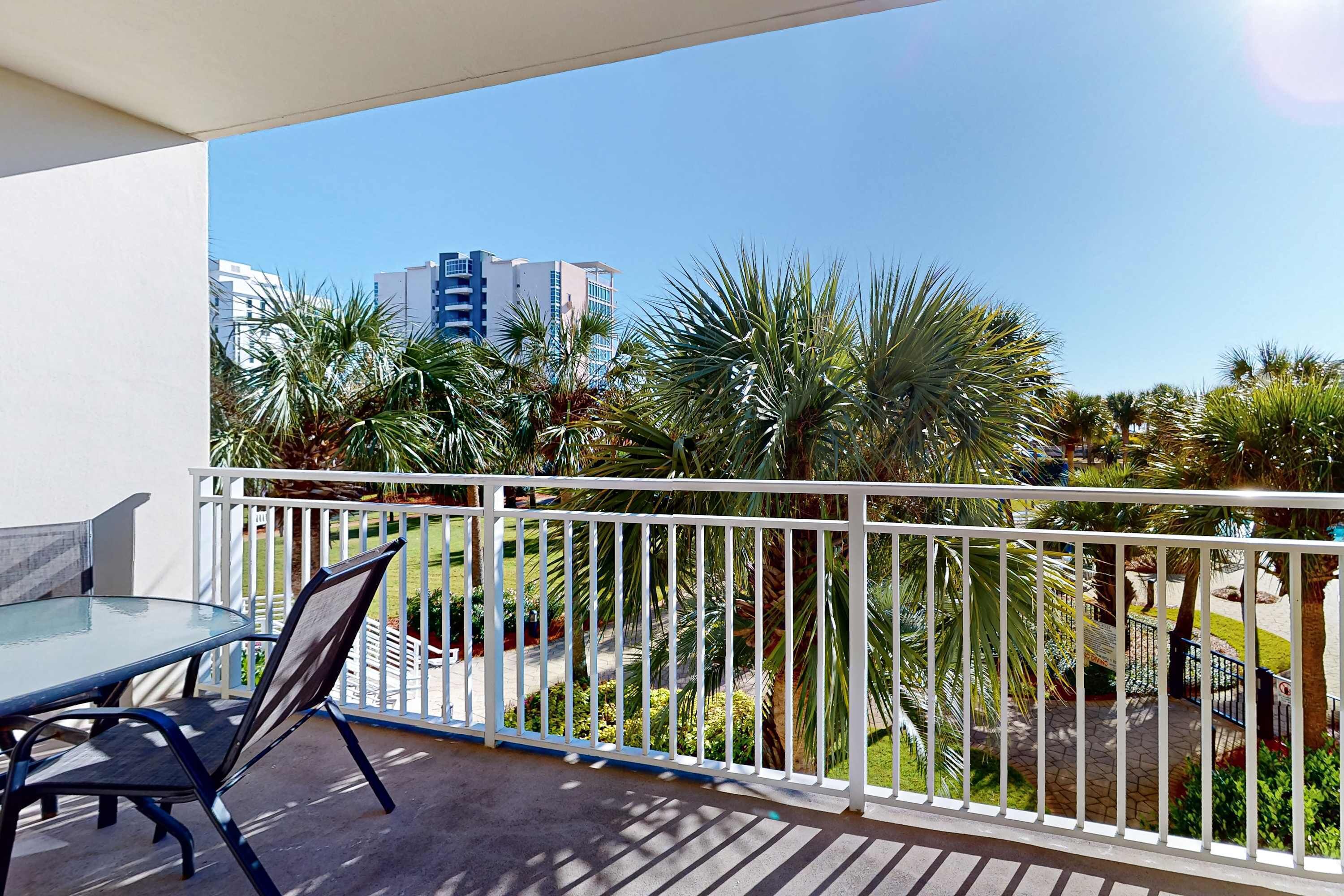 A balcony with a glass table and chairs, white railing, and palm trees in the background.