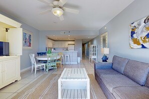 A modern living room with a grey sofa, a white coffee table, and a dining area with a white table and chairs. - Beautiful, waterfront condo w two shared pools, gym, & easy beach access (Destin)