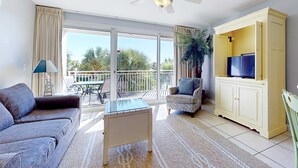 Una sala de estar con un sofá, una mesa de centro, un televisor sobre un mueble bajo y un balcón con vistas a palmeras.