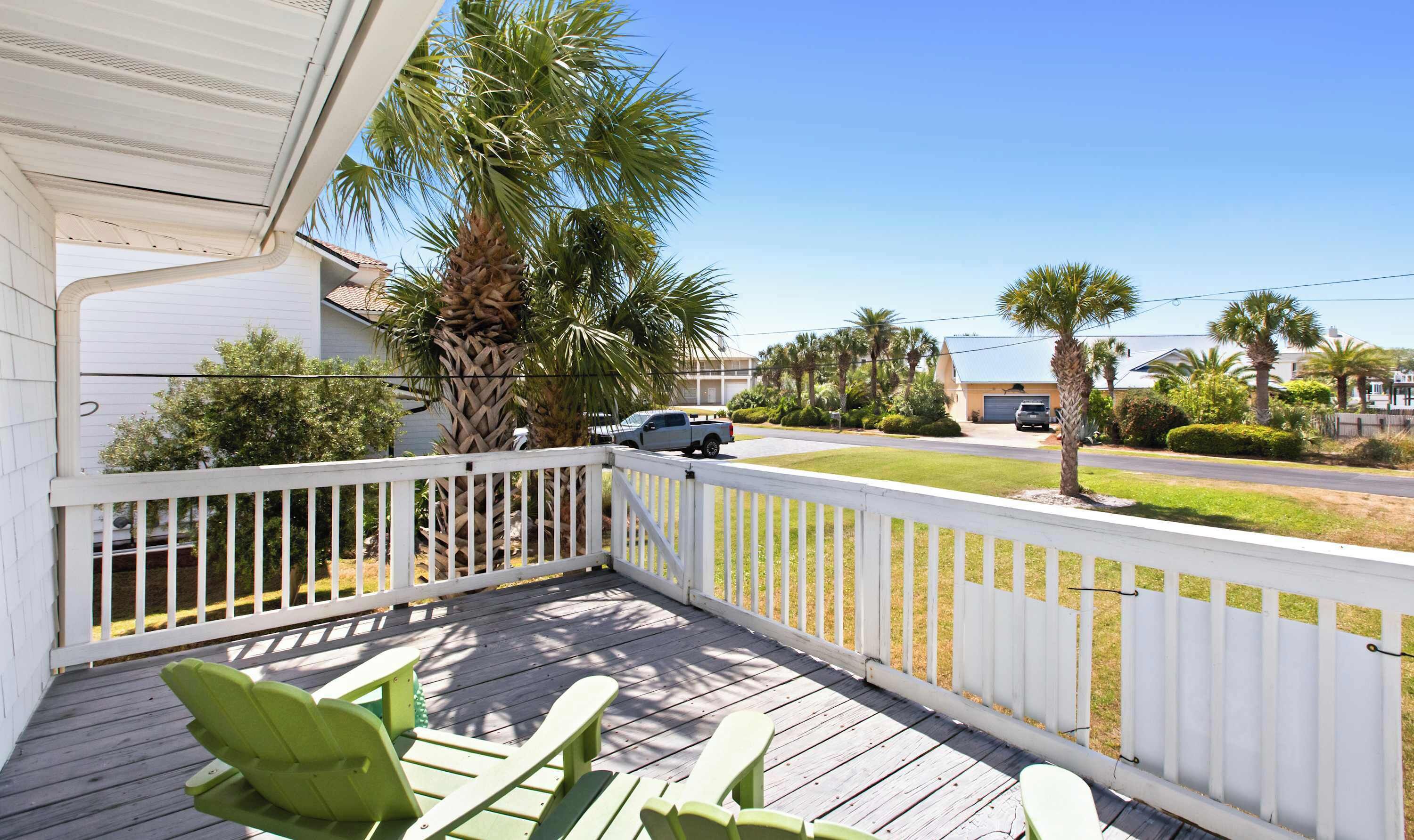 A wooden deck with chairs, a white fence, and a view of a street with houses and palm trees.