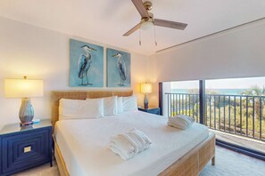 Une chambre d’hôtel avec un grand lit, deux tableaux représentant des oiseaux accrochés au mur, un ventilateur de plafond et un balcon avec vue sur la mer.