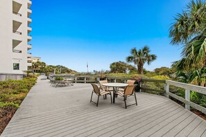 Une terrasse en bois avec une table et des chaises, entourée de verdure et avec un bâtiment en arrière-plan.