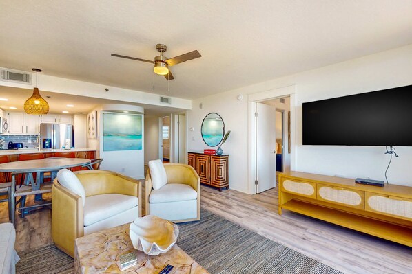 A modern living room with a flat-screen TV, two armchairs, a coffee table, a ceiling fan, and a dining area with a table and chairs. - Second floor oceanfront condo with pool access, ocean view, WiFi, W/D, and AC (Santa Rosa Beach)