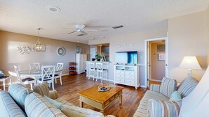 A spacious living room with a large sofa, a coffee table, and a TV. There is a dining area with a table and chairs, and a kitchen in the background. - Gorgeous beachfront condo with shared pool, hot tub and beach access (Panama City Beach)
