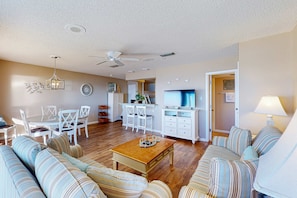 A spacious living room with a large sofa, a coffee table, and a TV. There is a dining area with a table and chairs, and a kitchen in the background. - Gorgeous beachfront condo with shared pool, hot tub and beach access (Panama City Beach)
