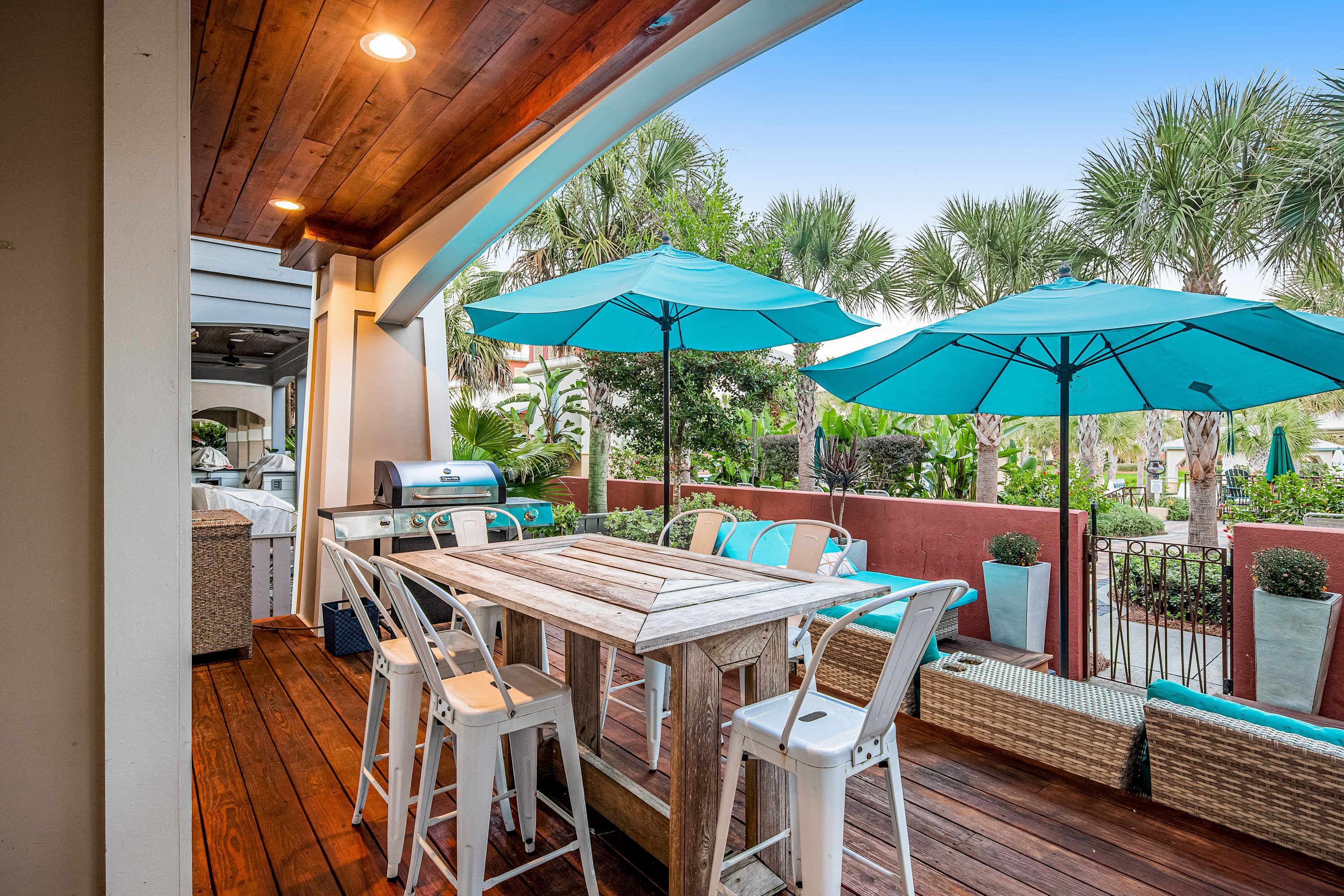 An outdoor patio with a wooden table and chairs, two blue umbrellas, and a grill.