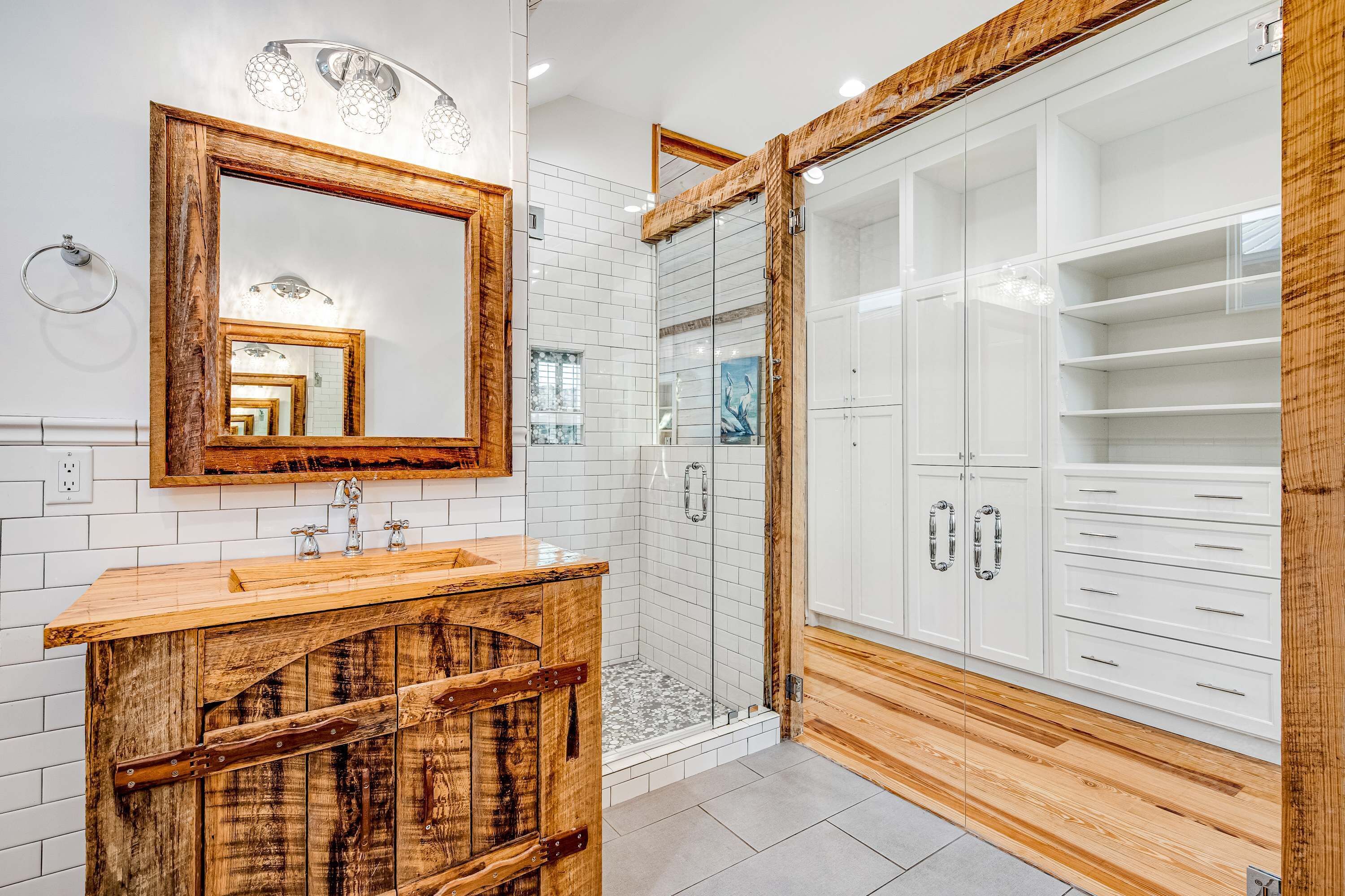 A modern bathroom with a wooden vanity, a large mirror, a walk-in shower, and a white storage cabinet.