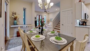 Un comedor con una mesa larga dispuesta para seis personas, una araña y vistas al salón y la cocina al fondo.