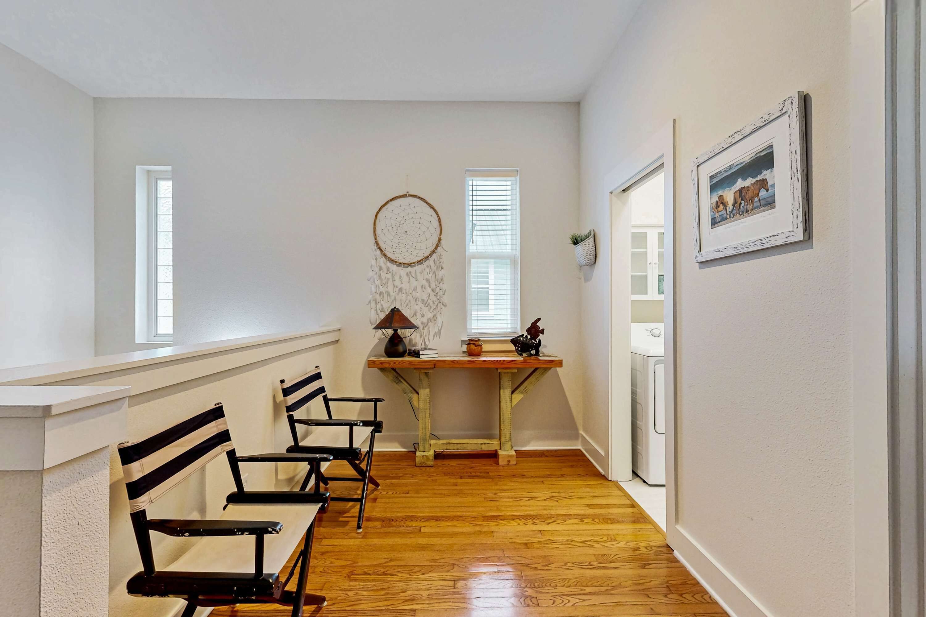 A hallway with wooden flooring, a white wall, a round dreamcatcher, a small table with a lamp, and a framed picture on the wall.