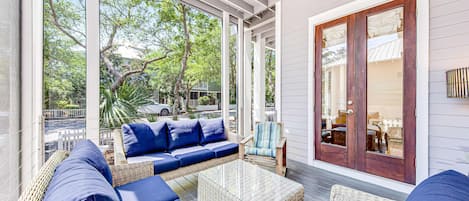 A covered porch with wicker furniture, a glass coffee table, and a view of the street and trees.