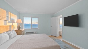 A bedroom with a large bed, a flat-screen TV mounted on the wall, a window with a view of the sea, and a door leading to another room. - Coastal Living Luxury Oceanfront Condo, Pool Beach (Virginia Beach)
