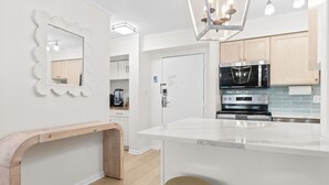 A modern kitchen with white cabinetry, a central island with stools, a built-in oven, and a hanging light fixture. - Coastal Living Luxury Oceanfront Condo, Pool Beach (Virginia Beach)