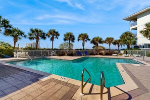 A swimming pool with a tiled deck, palm trees, and a white building in the background. - Cozy coastal condo with shared pool and easy beach access (Navarre)