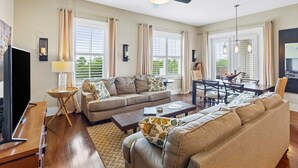 Ein geräumiges Wohnzimmer mit Sofa, Couchtisch und Fernsehtisch. Große Fenster mit Vorhängen, ein Deckenventilator und ein Essbereich mit Stühlen und Tisch.
