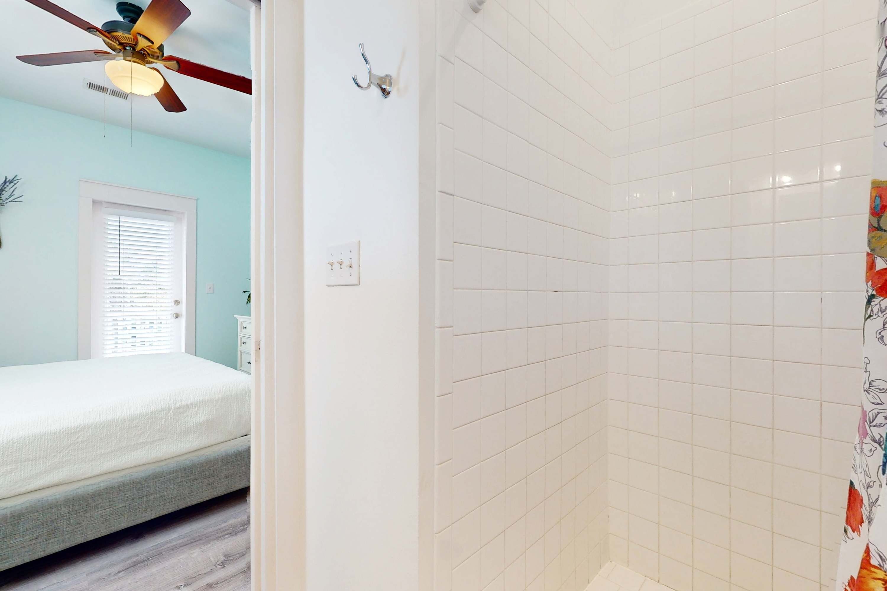 A bedroom with a bed, a ceiling fan, a window with blinds, and a bathroom with white tiles and a shower curtain.