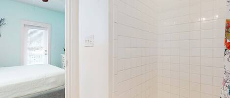 A bedroom with a bed, a ceiling fan, a window with blinds, and a bathroom with white tiles and a shower curtain.