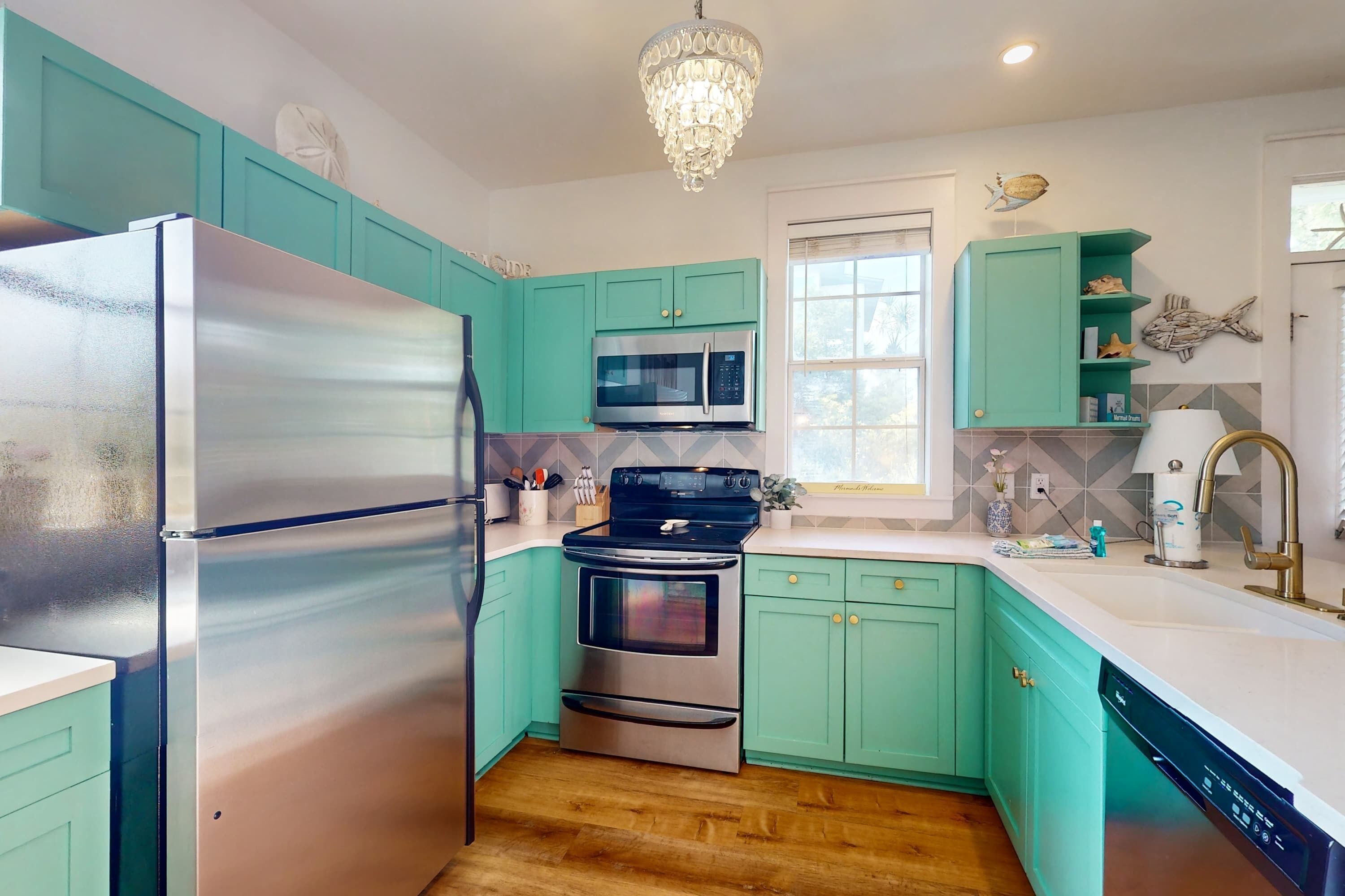 A kitchen with teal cabinets, stainless steel appliances, a chandelier, and a window with a view.