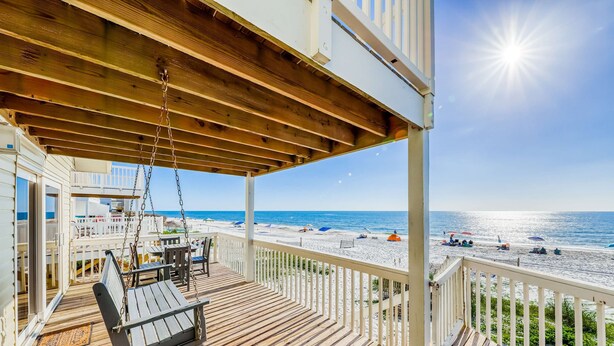 Eine Holzterrasse mit Schaukelstuhl, Tisch und Stühlen mit Blick auf einen Strand, der mit Sonnenschirmen und Liegestühlen ausgestattet ist.
