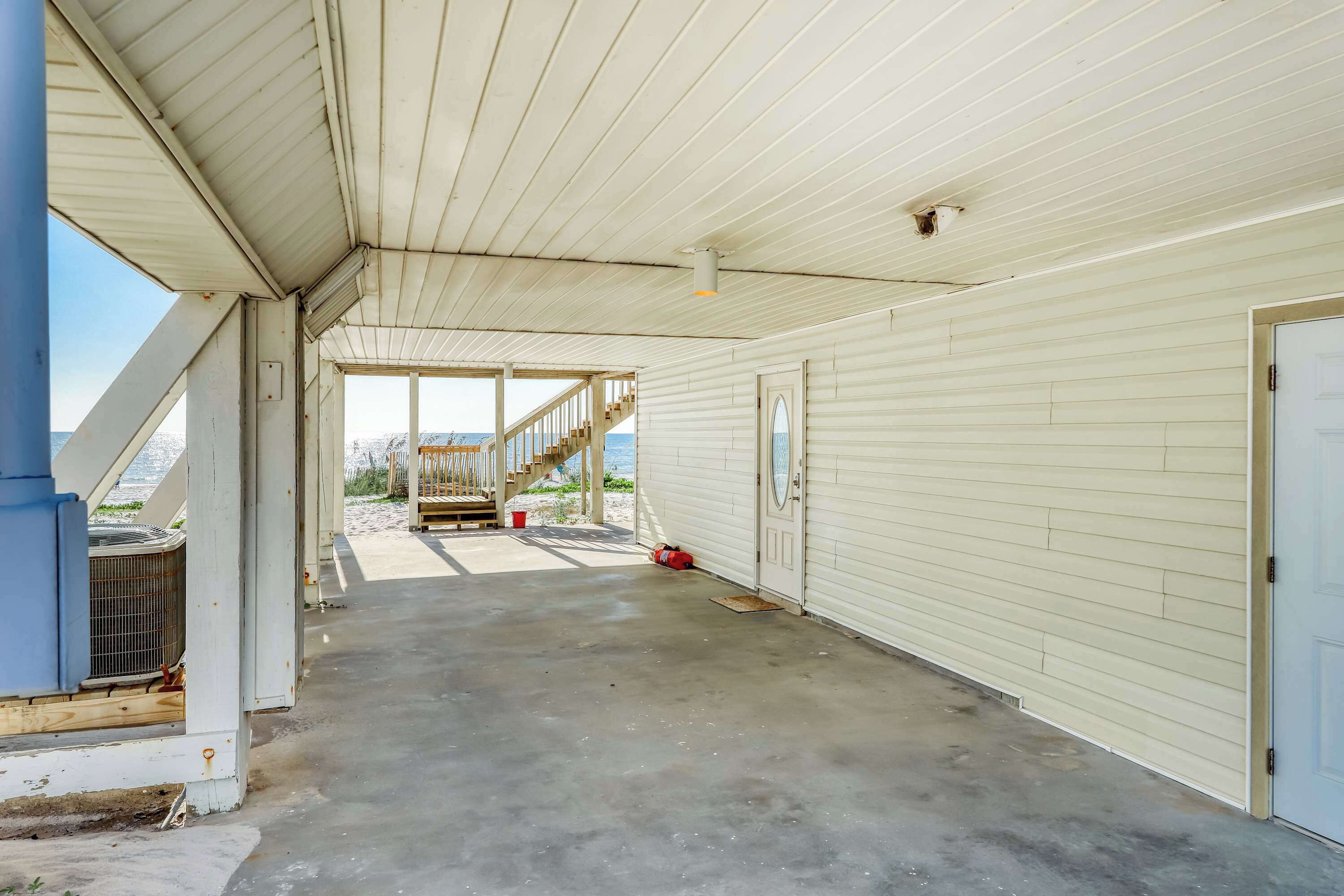 Un espace extérieur abrité, doté d’un sol en béton, d’un escalier en bois menant à la plage et d’une porte située à droite.