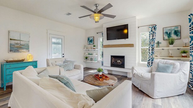 Una sala de estar con un sofá blanco, un sillón blanco, un aparador verde azulado, una chimenea, un televisor montado y un ventilador de techo.