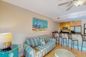 Ein Wohnzimmer mit einem gestreiften Sofa, einem Glastisch, einem Deckenventilator und einer Küchenzeile mit Kühlschrank und Barhocker.