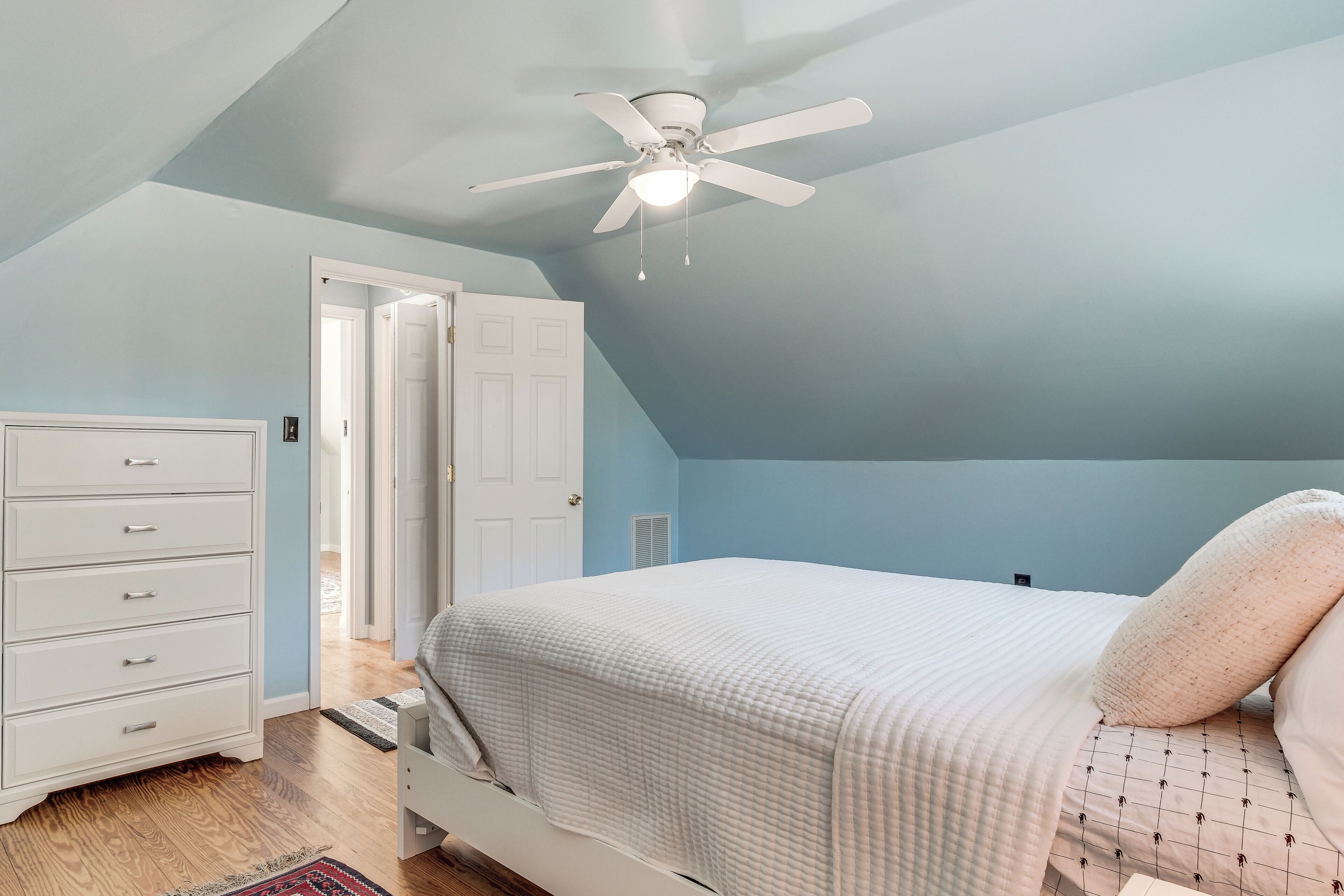 A bedroom with a bed, a dresser, a ceiling fan, and a door leading to another room.
