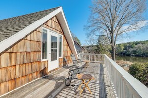 Um deck de madeira com móveis de exterior, guarda-corpo branco e vista para um lago.