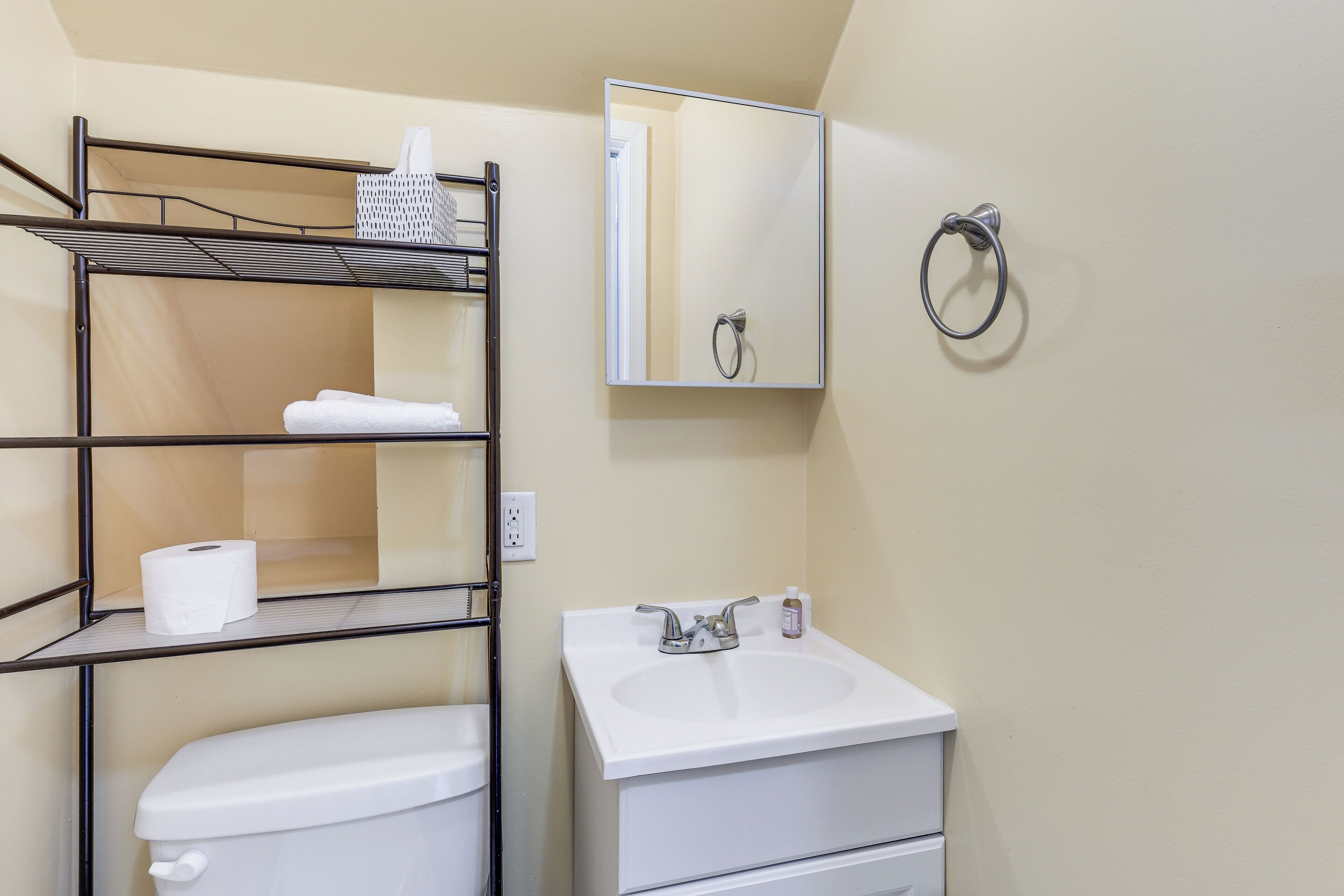 A bathroom with a white toilet, a sink with a mirror above it, and a metal shelf unit with towels and a roll of toilet paper.