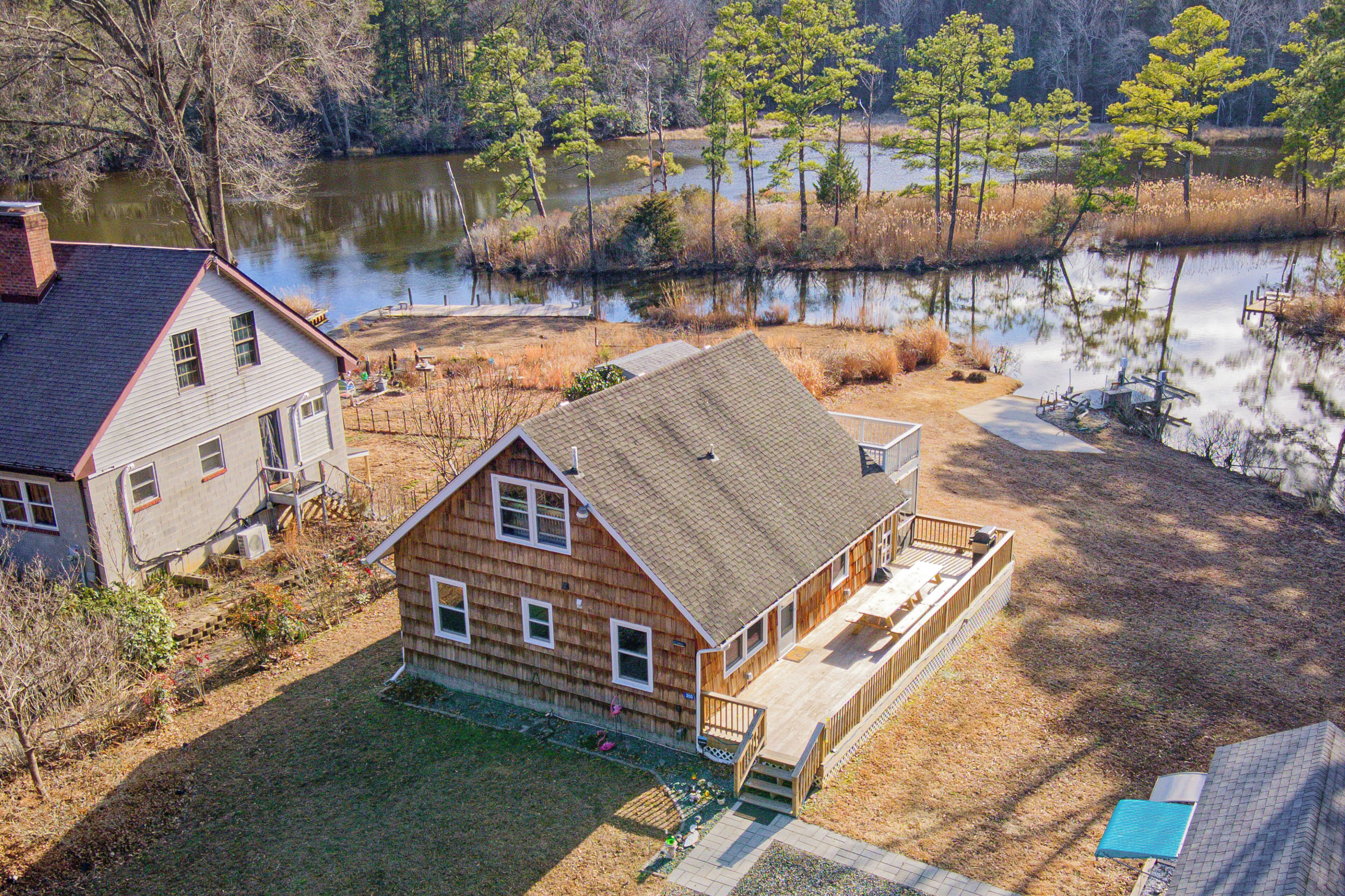 A two-story wooden house with a deck, a pond in the background, and a grassy yard.