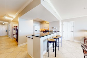 A modern kitchen with a central island, bar stools, and a view into the adjacent dining area. - Beachfront 16th-floor condo with Gulf views, pool, game room, balcony, gym, W&D (Panama City Beach)