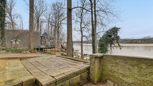 Une terrasse en pierre surplombant un lac paisible, entourée d’arbres et d’une maison avec un quai.