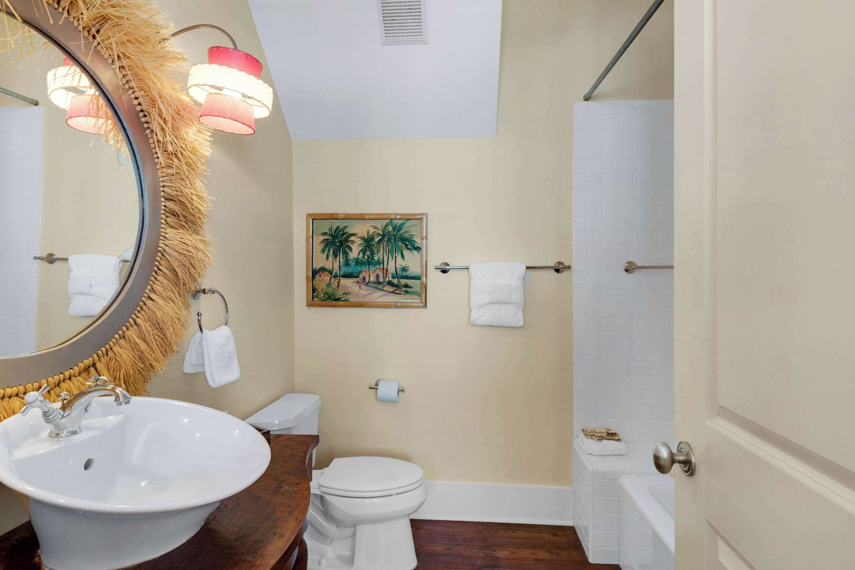 A bathroom with a round mirror, white sink, toilet, and a painting of palm trees on the wall.