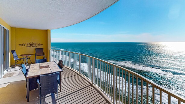 Ein Balkon mit Holztisch und -stühlen mit Blick auf einen Strand, Wellen und einen klaren Himmel.