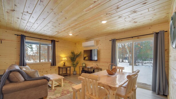 Interior de una cabaña de madera con sofá, mesa de comedor, sillas y televisor montado en la pared.