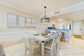 A dining area with a table and chairs, a hanging light fixture, and a kitchen in the background. - Gorgeous beach view condo, Shared pool and Beach access (Panama City Beach)