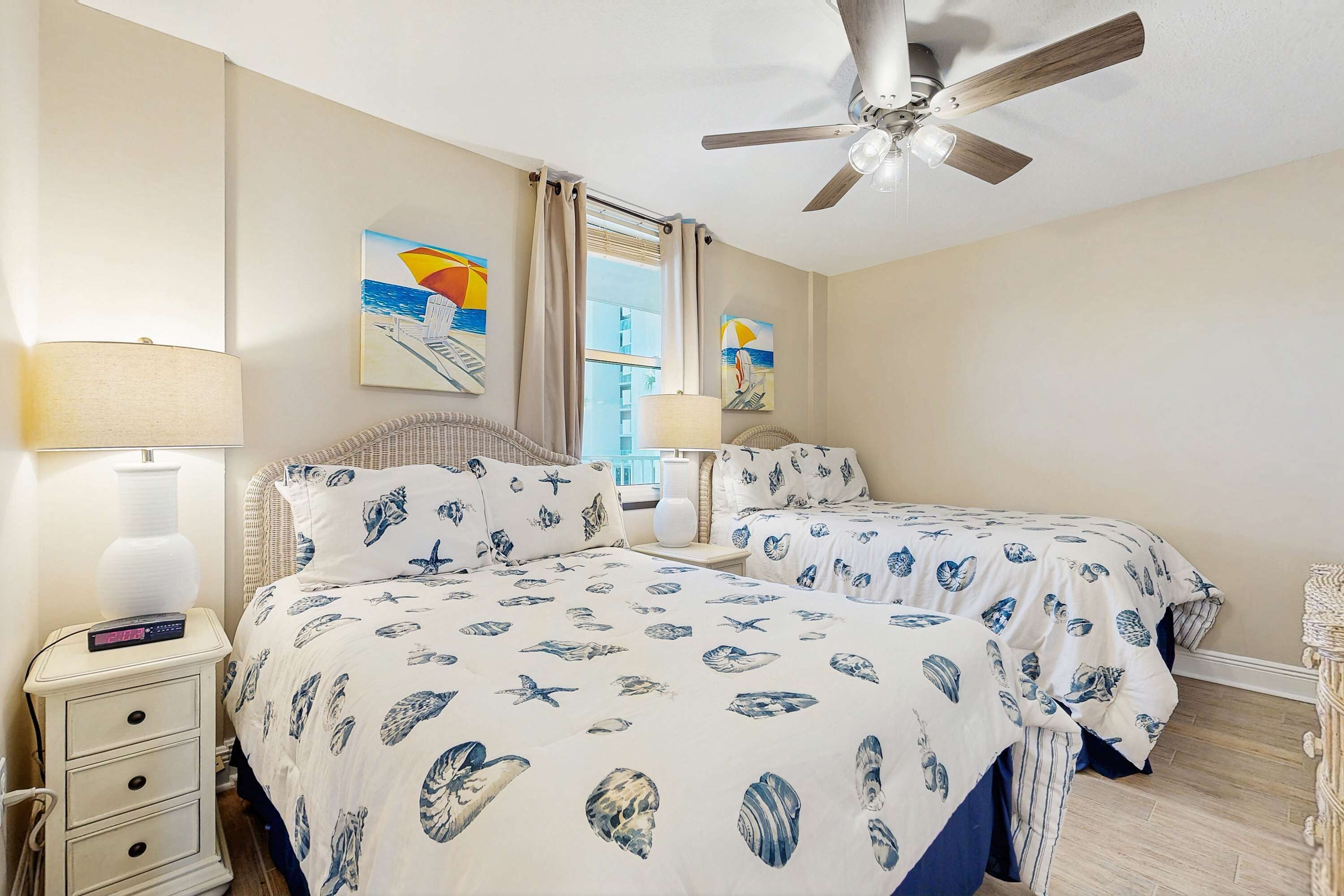 A hotel room with two beds, a ceiling fan, a nightstand with a lamp, and artwork on the walls.