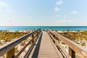 Ein hölzerner Steg führt zu einem Strand mit Sonnenschirmen und badenden Gästen.