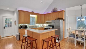 A kitchen with wooden cabinets, a central island with stools, a refrigerator, and a dining area with a table and chairs. - Centrally located home with enclosed yard, grill, high ceilings, smart TVs, & AC (Santa Rosa Beach)