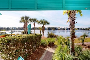 A serene lakeside area with a paved walkway, palm trees, and a view of houses across the water. - Lakefront dog-friendly condo with water views, pool, hot tub & beach access (Panama City Beach)