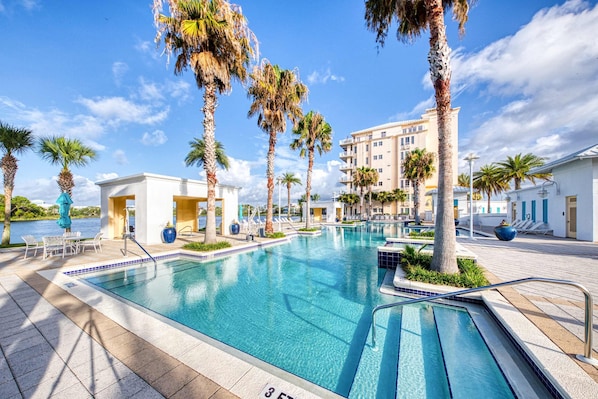 A swimming pool with clear blue water, surrounded by palm trees and a building with balconies. - Lakefront dog-friendly condo with water views, pool, hot tub & beach access (Panama City Beach)