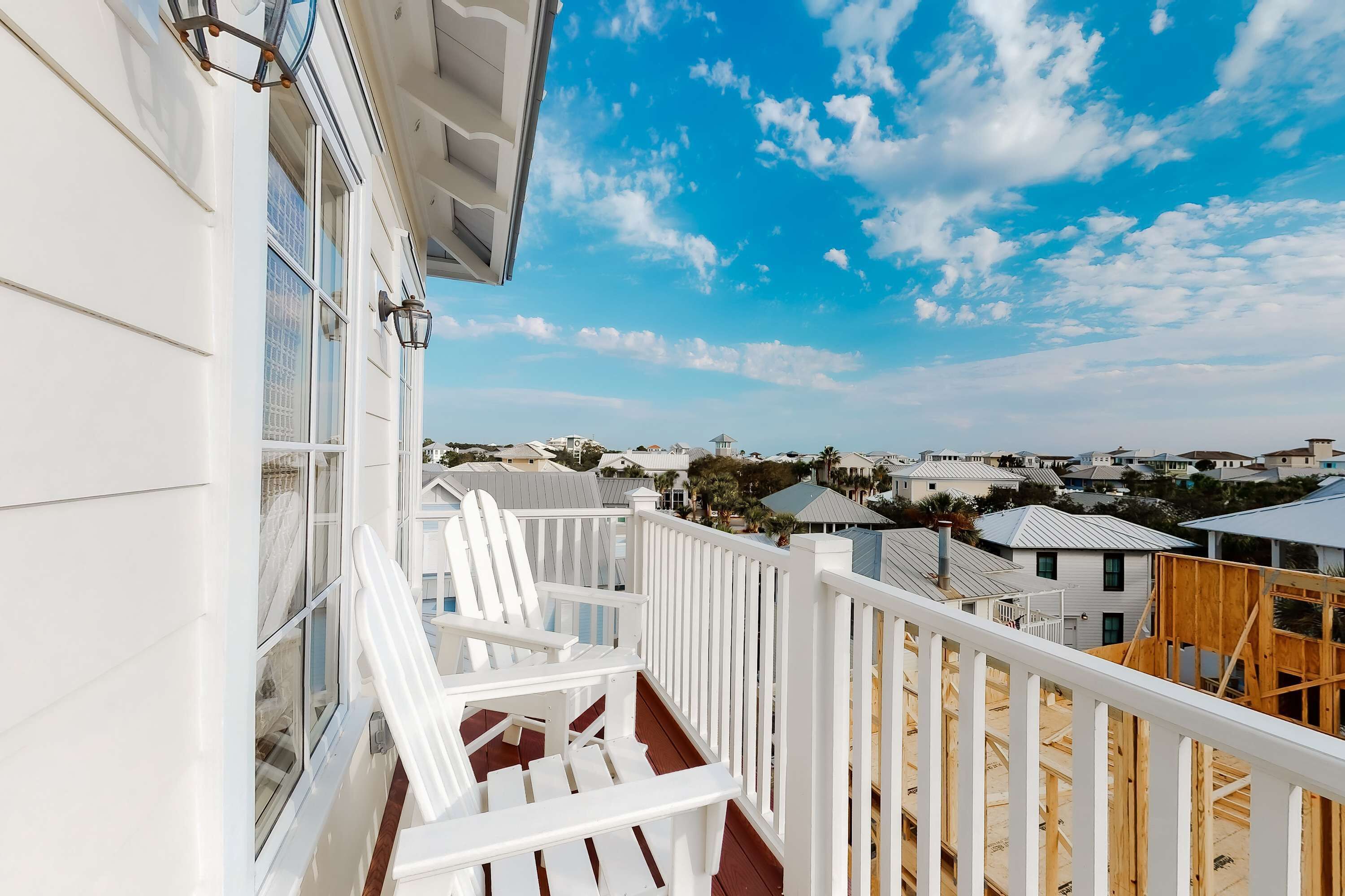 Ein Balkon mit weißen Geländern und Stühlen, der auf einen Wohnbereich mit Häusern und einem klaren, blauen Himmel blickt.