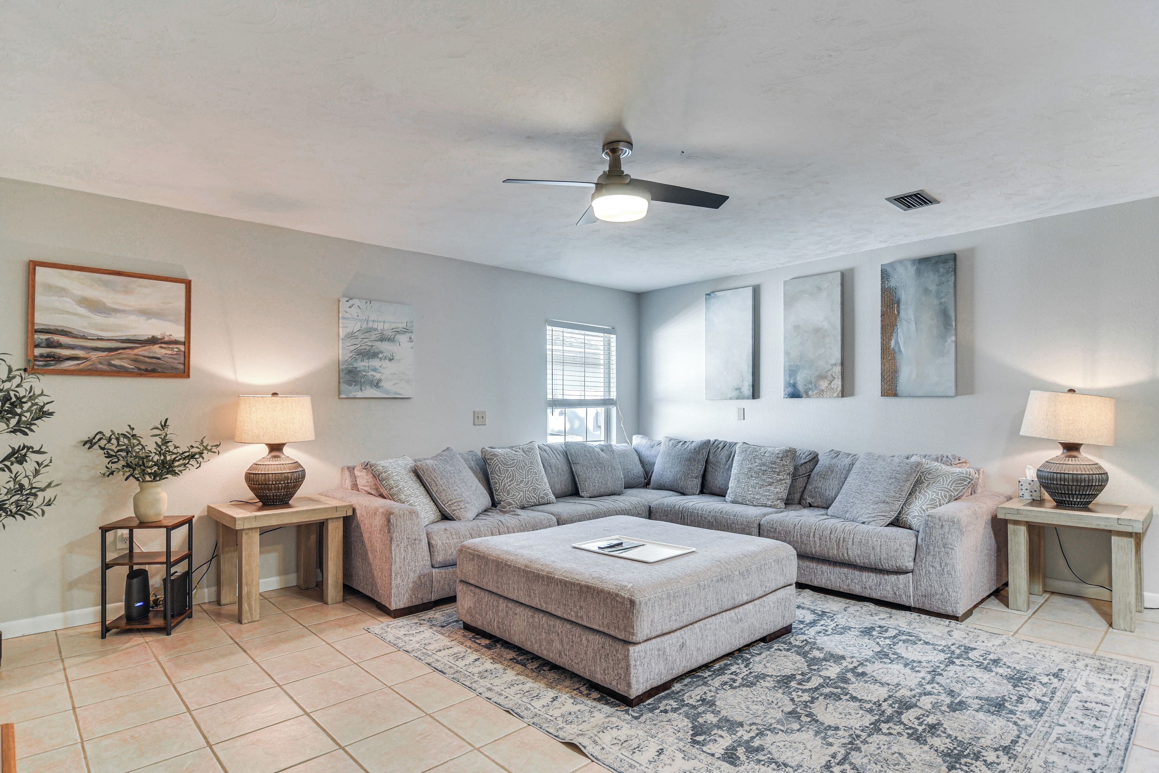 A spacious living room with a large L-shaped sectional sofa, a matching ottoman, two side tables with lamps, a ceiling fan, and artwork on the walls.