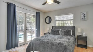 Une chambre avec un lit, un ventilateur de plafond, une table de chevet, une fenêtre avec des stores et une vue sur une piscine à l’extérieur.